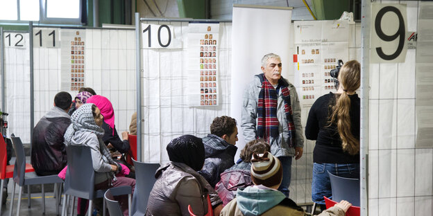 Menschen stehen und sitzen an numerierten Ständen mit Trennwänden Menschen stehen und sitzen an numerierten Ständen mit Trennwänden