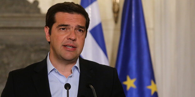Ein Mann mit dunklen Haaren spricht in ein Mikrofon, hinter ihm die griechische und die europäische Flagge Ein Mann mit dunklen Haaren spricht in ein Mikrofon, hinter ihm die griechische und die europäische Flagge
