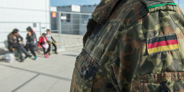 Die Uniform eines Bundeswehrsoldaten, im Hintergrund eine kleine Gruppe von Menschen Die Uniform eines Bundeswehrsoldaten, im Hintergrund eine kleine Gruppe von Menschen