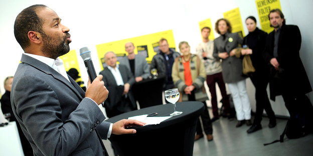Markus Beeko steht an einem Tisch und spricht in ein Mikrofon, im Hintergrund Zuhörer