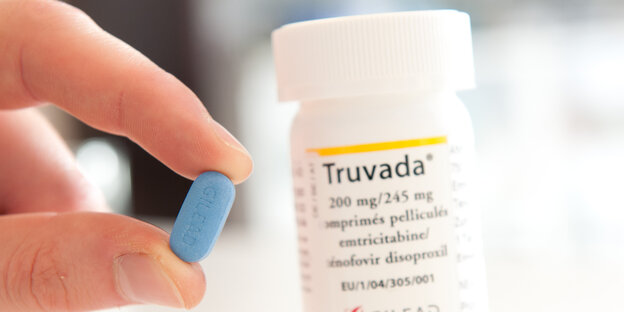 Zwei Finger halten eine kleine blaue Pille, dahinter die Pillendose mit Aufschrift „Truvada“ Zwei Finger halten eine kleine blaue Pille, dahinter die Pillendose mit Aufschrift „Truvada“