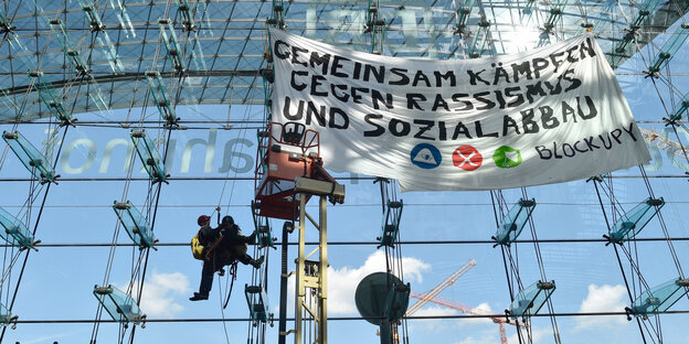 eine Glasfassade, vor der sich Menschen abseilen, und ein Plakat mit der Aufschrift „Gemeinsam kämpfen gegen Rassismus und Sozialabbau“