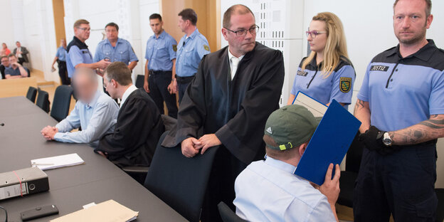 Mehrere Menschen, darunter Anwälte und Polizisten, stehen in einem Gerichtssaal Mehrere Menschen, darunter Anwälte und Polizisten, stehen in einem Gerichtssaal