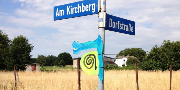 An einem Straßenschild vor einem Feld hängen bunte Kinderbasteleien