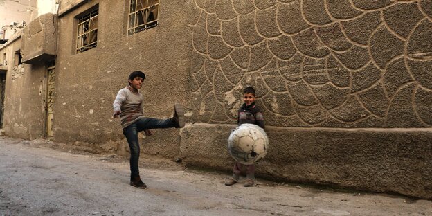 Zwei Kinder in einer Straße, einer schießt einen Fußball