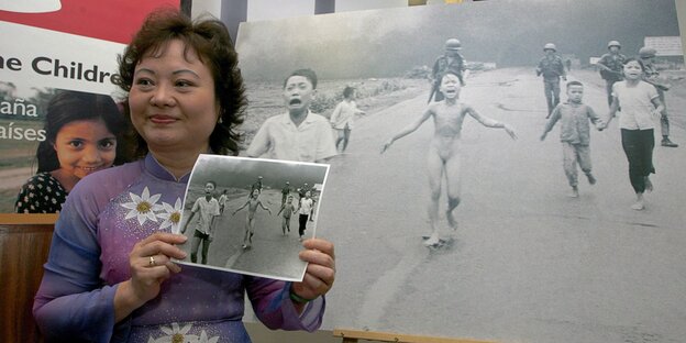 Eine Frau steht neben einem Foto, das ein vor Napalm fliehendes nacktes Mädchen im Vitenamkrieg zeigt Eine Frau steht neben einem Foto, das ein vor Napalm fliehendes nacktes Mädchen im Vitenamkrieg zeigt