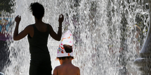 Eine Frau an einem Springbrunnen dreht ihrem Kind den Rücken zu. Das Kind sitzt am Springbrunnenrand mit einem Eimer Eine Frau an einem Springbrunnen dreht ihrem Kind den Rücken zu. Das Kind sitzt am Springbrunnenrand mit einem Eimer