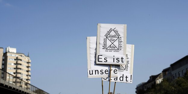 Vor blauem Himmel: Schilder mit der Aufschrift „Es ist unsere Stadt“, Häuserzeilen rechts und links am Rand Vor blauem Himmel: Schilder mit der Aufschrift „Es ist unsere Stadt“, Häuserzeilen rechts und links am Rand