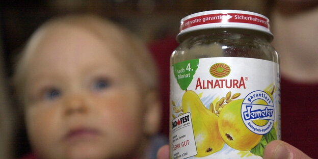 Ein Glas mit Babynahrung von Alnatura, im Hintergrund das Gesicht eines Kleinkinds Ein Glas mit Babynahrung von Alnatura, im Hintergrund das Gesicht eines Kleinkinds