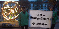Ein brennendes Ceta-Symbol. Davor stehen zwei Frauen mit einem Plakat auf dem steht: Ceta – brandgefährlich. Darunter prangt der Greenpeace-Schriftzug