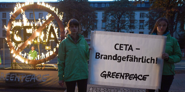 Ein brennendes Ceta-Symbol. Davor stehen zwei Frauen mit einem Plakat auf dem steht: Ceta – brandgefährlich. Darunter prangt der Greenpeace-Schriftzug Ein brennendes Ceta-Symbol. Davor stehen zwei Frauen mit einem Plakat auf dem steht: Ceta – brandgefährlich. Darunter prangt der Greenpeace-Schriftzug
