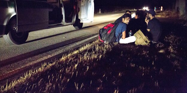 Flüchtlinge sitzen im August 2015 am Straßenrand, es ist dunkel, ein großes Auto steht daneben