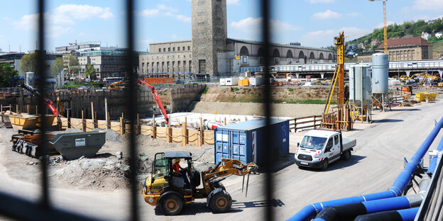 Durch Gitter blickt man auf eine Baustelle, auf der ein Bagger fährt Durch Gitter blickt man auf eine Baustelle, auf der ein Bagger fährt
