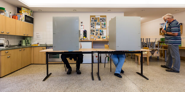 In einem Wahllokal, zwei Kabinen, dahinter zwei Menschen, deren Beine man sieht In einem Wahllokal, zwei Kabinen, dahinter zwei Menschen, deren Beine man sieht