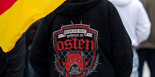 Auf einem schwarzem Pullover steht „Natürliche Härte geboren im Osten“, daneben hängt eine Deutschlandfahne Auf einem schwarzem Pullover steht „Natürliche Härte geboren im Osten“, daneben hängt eine Deutschlandfahne