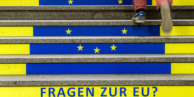 Ein Manh geht eine Treppe hoch, auf der die Europaflagge und die Aufschrift "Fragen zur EU?" zu sehen sind Ein Manh geht eine Treppe hoch, auf der die Europaflagge und die Aufschrift "Fragen zur EU?" zu sehen sind