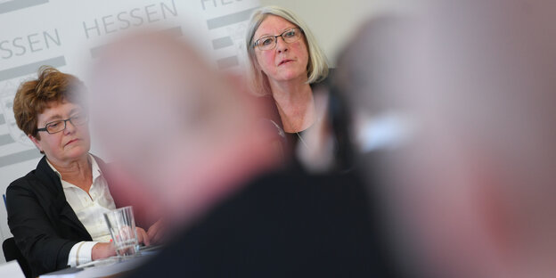 Zwei Frauen sitzen an einem Tisch, man sieht sie von vorne, Männer, von denen die Hinterköpfe unscharf zu sehen sind, sitzen ihnen gegenüber Zwei Frauen sitzen an einem Tisch, man sieht sie von vorne, Männer, von denen die Hinterköpfe unscharf zu sehen sind, sitzen ihnen gegenüber
