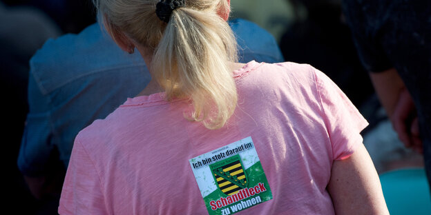Frau von hinten, auf deren rosa T-Shirt steht: "Ich bin stolz darauf, im Schandfleck zu leben" Frau von hinten, auf deren rosa T-Shirt steht: "Ich bin stolz darauf, im Schandfleck zu leben"