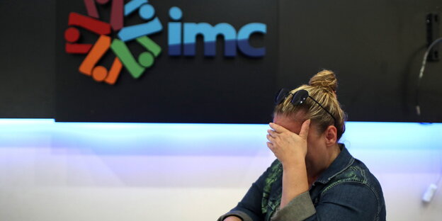 Eine Frau hält sich mit gesenktem Kopf die Hand vor die Augen, hinter ihr das IMC-Logo Eine Frau hält sich mit gesenktem Kopf die Hand vor die Augen, hinter ihr das IMC-Logo