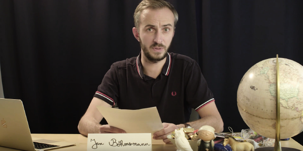 Jan Böhmermann sitzt am Tisch, neben sich ein Globus. In der Hand hält er ein Blatt Papier