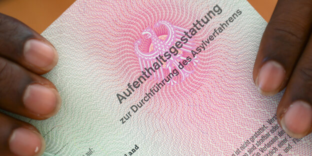 Eine Person hält ein Dokument mit der Aufschrift „Aufenthaltsgestattung“ Eine Person hält ein Dokument mit der Aufschrift „Aufenthaltsgestattung“