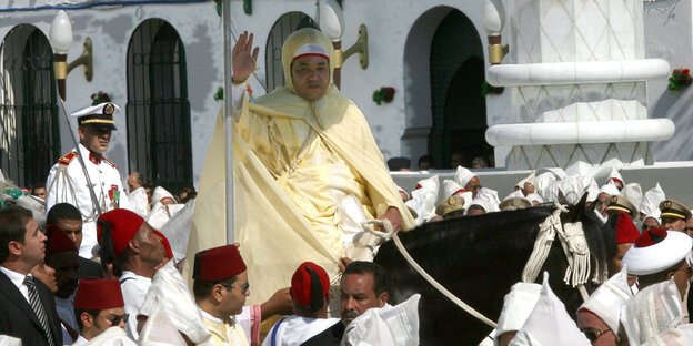 Mohammed VI. reitet auf einem Pferd unter einem roten Sonnenschirm durch eine Menschenmenge Mohammed VI. reitet auf einem Pferd unter einem roten Sonnenschirm durch eine Menschenmenge