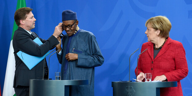 Steffen Seibert setzt Nigerias Präsident Buhari Kopfhörer auf während Angela Merkel daneben steht Steffen Seibert setzt Nigerias Präsident Buhari Kopfhörer auf während Angela Merkel daneben steht