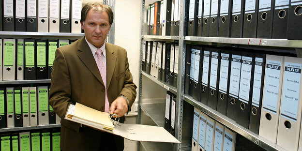Ein Mann mit einem Aktenordner, im Hintergrund Regale mit weiteren Aktenordnern Ein Mann mit einem Aktenordner, im Hintergrund Regale mit weiteren Aktenordnern