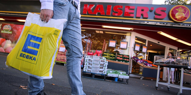 Vor einem Supermarkt mit der Aufschrift Kaiser's geht jemand mit einer gelben Edeka-Tüte vorbei Vor einem Supermarkt mit der Aufschrift Kaiser's geht jemand mit einer gelben Edeka-Tüte vorbei