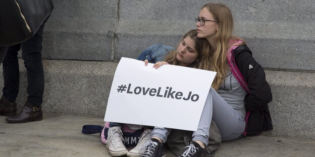 Zwei Mädchen sitzen aneinander gelehnt und halten ein Schild, auf dem „#LoveLikeJo“ steht Zwei Mädchen sitzen aneinander gelehnt und halten ein Schild, auf dem „#LoveLikeJo“ steht