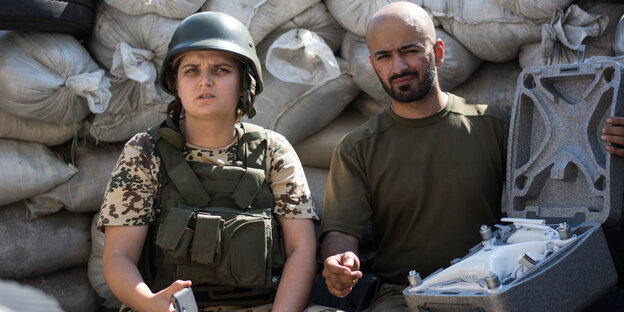 Eine Frau in kugelsicherer Weste und mit Helm auf dem Kopf sitzt neben einem Mann mit kahlem Kopf und Bart. Sie sitzen vor aufgestapelten Sandsäcken