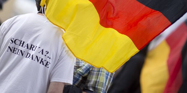 Ein Mann von hinten mit Deutschlandfahne, auf seinem Shirt steht „Scharia nein danke“ Ein Mann von hinten mit Deutschlandfahne, auf seinem Shirt steht „Scharia nein danke“