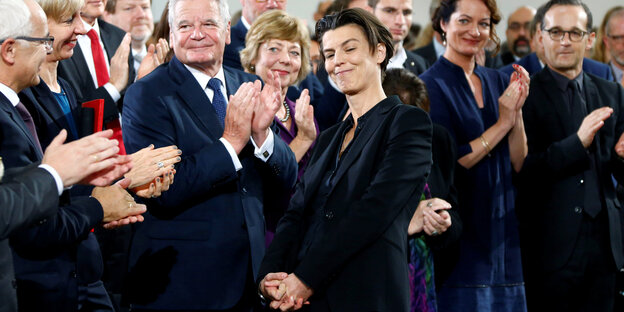 Eine lächelnde Frau im Vordergrund, dahinter applaudierend das Publikum Eine lächelnde Frau im Vordergrund, dahinter applaudierend das Publikum