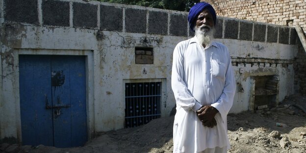 Ein Mann in weißer Kleidung und einem blauen Turban steht vor einem weißen Gebäude Ein Mann in weißer Kleidung und einem blauen Turban steht vor einem weißen Gebäude