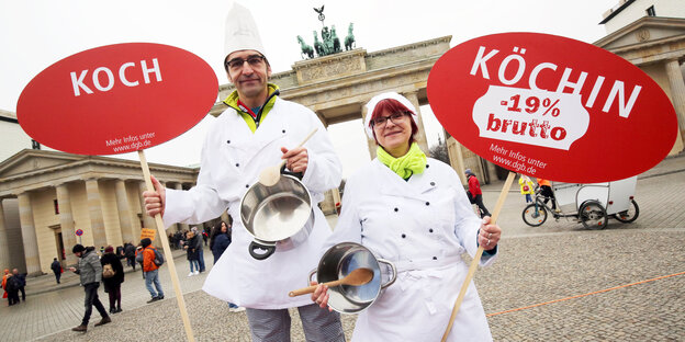 Ein Koch und eine Köchin in Arbeitskleidung halten vor dem Brandenburger Tor Schilder mit der Aufschrift „Koch“ und „Köchin -19% brutto“ in die Luft Ein Koch und eine Köchin in Arbeitskleidung halten vor dem Brandenburger Tor Schilder mit der Aufschrift „Koch“ und „Köchin -19% brutto“ in die Luft