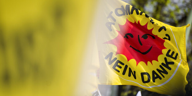 Im Vordergrund eine gelbe verschwommene Flagge, im Hintergrund auch eine gelbe Flagge mit einer roten Sonne und der Aufschrift: Atomkraft, Nein Danke. Im Vordergrund eine gelbe verschwommene Flagge, im Hintergrund auch eine gelbe Flagge mit einer roten Sonne und der Aufschrift: Atomkraft, Nein Danke.