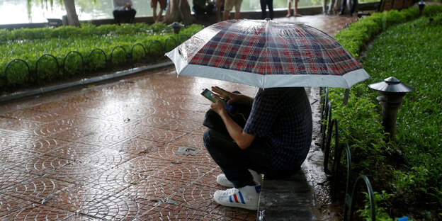 Ein Mann hockt mit einem Regenschirm im Park und spielt auf seinem Smartphone Ein Mann hockt mit einem Regenschirm im Park und spielt auf seinem Smartphone