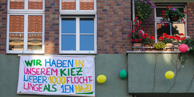 An einer grünen Hauswand hängt ein Transparent „Wir haben in unserem Kiez lieber 1000 Flüchtlinge, als 1en Nazi“. Daneben ein Balkon mit bunten Blumen An einer grünen Hauswand hängt ein Transparent „Wir haben in unserem Kiez lieber 1000 Flüchtlinge, als 1en Nazi“. Daneben ein Balkon mit bunten Blumen