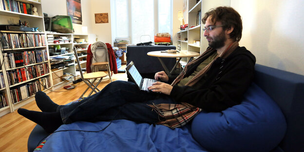 Ein junger Mann sitzt mit ausgestreckten Beinen auf einer Couch und schreibt auf einem Laptop Ein junger Mann sitzt mit ausgestreckten Beinen auf einer Couch und schreibt auf einem Laptop