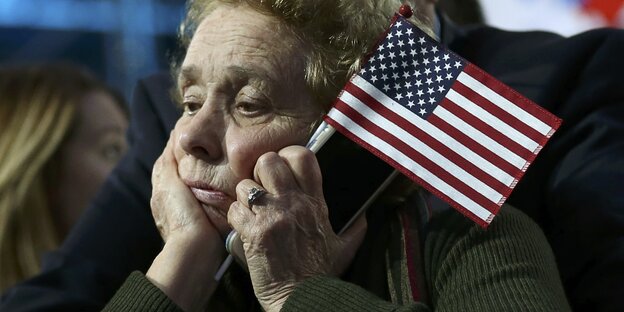 Eine enttäuscht schauende ältere Dame hält eine Amerikaflagge und ein Smartphone in der linken Hand. Sie stützt den Kopf auf die rechte Hand. Eine enttäuscht schauende ältere Dame hält eine Amerikaflagge und ein Smartphone in der linken Hand. Sie stützt den Kopf auf die rechte Hand.