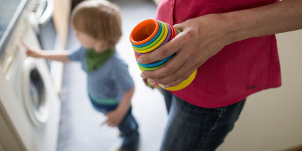 Ein Kind steht auf der Boden, seine Mutter hält ein Spielzeug in der Hand Ein Kind steht auf der Boden, seine Mutter hält ein Spielzeug in der Hand