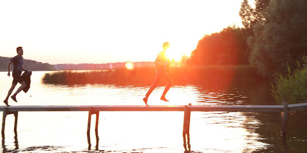 Zwei Männer rennen über einen Steg, der über Wasser führt, dahinter ein Sonnenuntergang Zwei Männer rennen über einen Steg, der über Wasser führt, dahinter ein Sonnenuntergang
