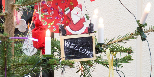 An einem Weihnachtsbaum hängt eine Tafel mit der Aufschrift „Welcome“, auf der ein hölzerner Weihnachtsmann sitzt An einem Weihnachtsbaum hängt eine Tafel mit der Aufschrift „Welcome“, auf der ein hölzerner Weihnachtsmann sitzt