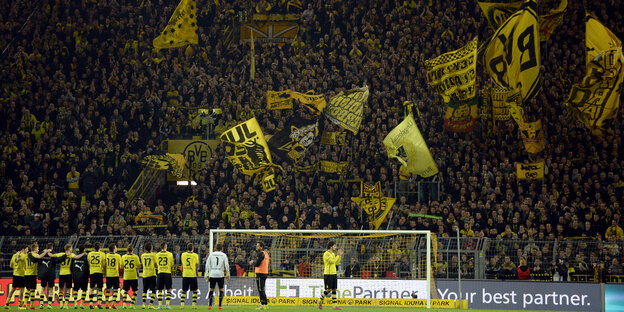 Ein Fußballteam in gelben Trickots steht vor der Fankurve eines Stadions, auf der gelbe Fahnen geschwenkt werden Ein Fußballteam in gelben Trickots steht vor der Fankurve eines Stadions, auf der gelbe Fahnen geschwenkt werden