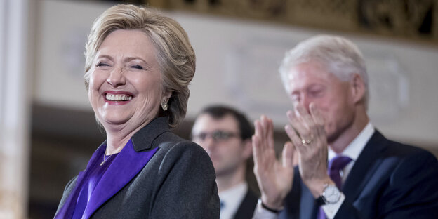Die demokratische US-Präsidentschaftskandidatin Hillary Clinton steht lachend an einem Rednerpult, hinter ihr klatscht ihr Ehemann Bill Clinton Die demokratische US-Präsidentschaftskandidatin Hillary Clinton steht lachend an einem Rednerpult, hinter ihr klatscht ihr Ehemann Bill Clinton