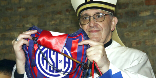 Ein Mann mit Brille und in Kardinalsgewand und -mütze hält einen Wimpel eines Fußballvereins hoch Ein Mann mit Brille und in Kardinalsgewand und -mütze hält einen Wimpel eines Fußballvereins hoch