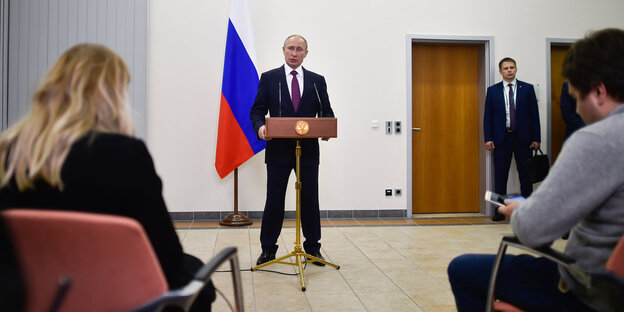 Im Hintergrund: Putin hinter einem Rednerpult, im Vordergrund: sitzende Journalisten, die ihm lauschen Im Hintergrund: Putin hinter einem Rednerpult, im Vordergrund: sitzende Journalisten, die ihm lauschen