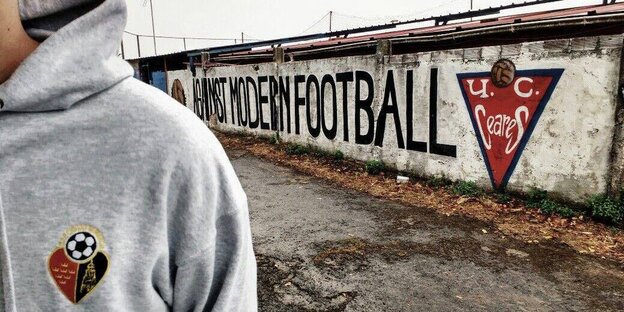 Im Vordergrund ein grauer Kapuzenpullover mit Vereinslogo, im Hintergrund steht auf einer Mauer "Against Modern Football" Im Vordergrund ein grauer Kapuzenpullover mit Vereinslogo, im Hintergrund steht auf einer Mauer "Against Modern Football"