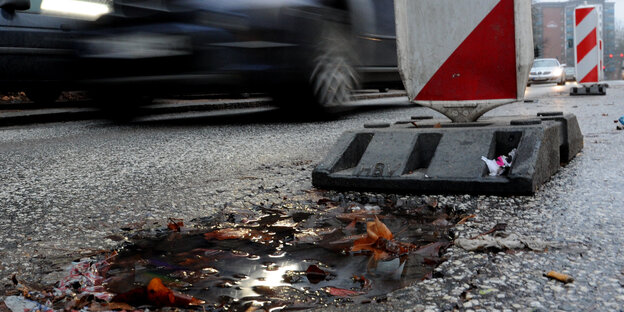 Schlagloch auf einer Straße, daneben eine Warnbake und vorbeifahrende Autos