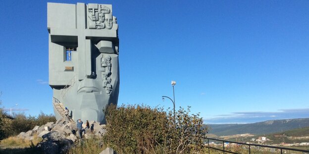 Das Denkmal einer Maske steht in karger Landschaft, darunter sind kleine Menschen zu sehen Das Denkmal einer Maske steht in karger Landschaft, darunter sind kleine Menschen zu sehen